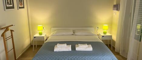 A neatly made bed with a blue quilt, two white pillows, and two folded towels on the bed. There are two bedside tables with lamps, a wooden hanger on the floor, and framed pictures on the wall.