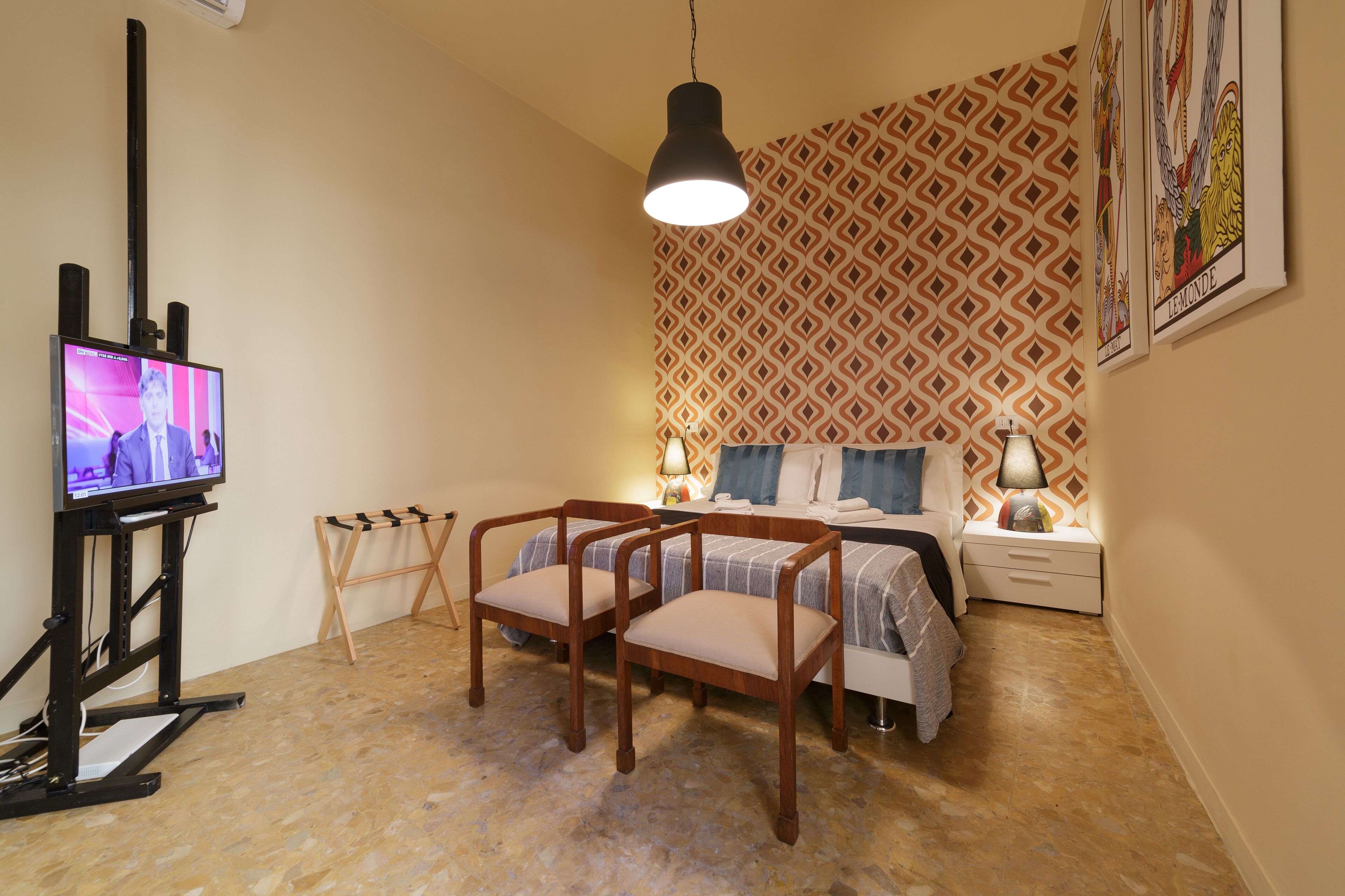Un dormitorio con una cama, dos sillas de madera, un televisor, una lámpara y un papel tapiz estampado.