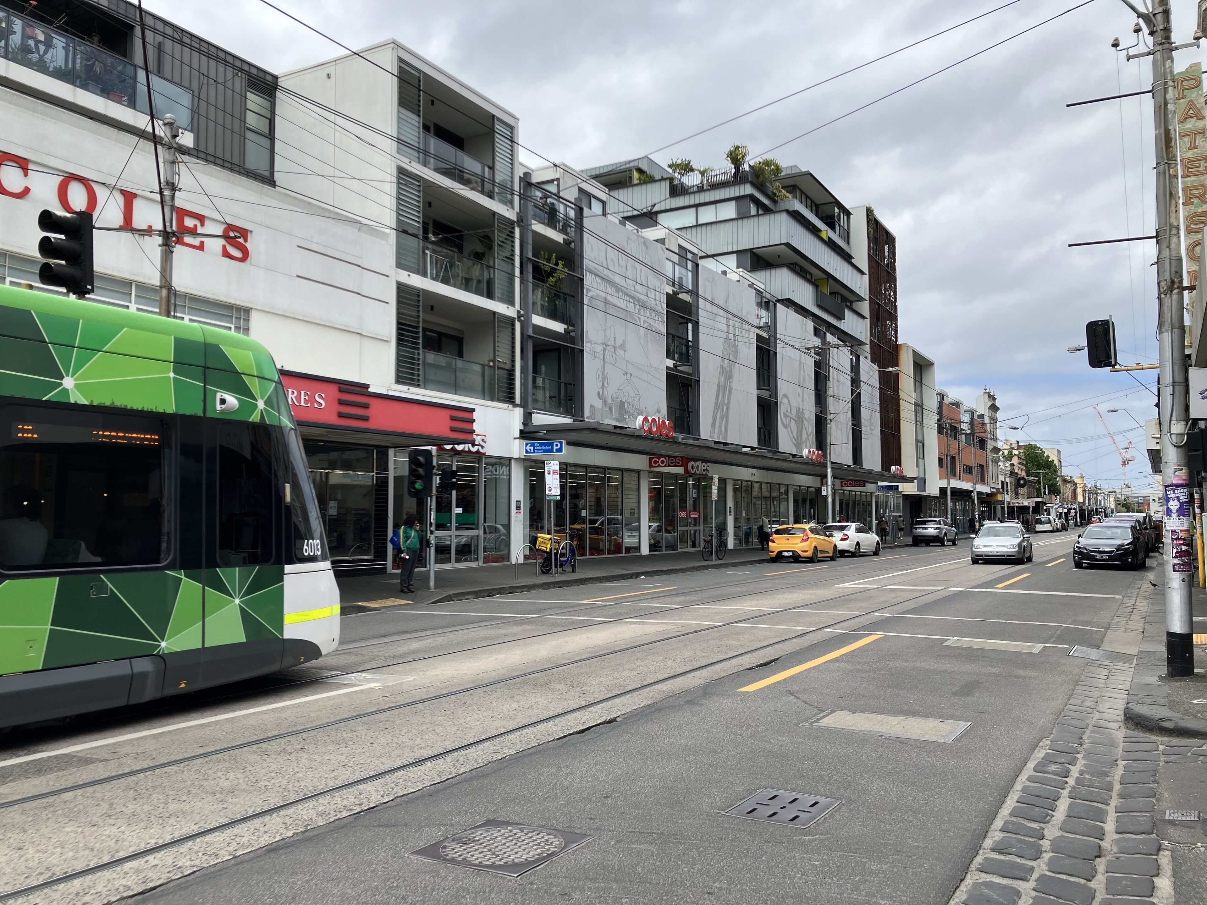 A tram passing by a modern building with a 'COLLES' sign, indicating a commercial area with shops.