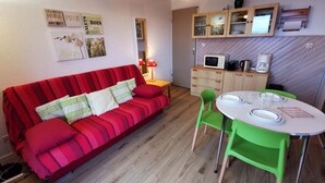 Een knus woongedeelte met een rode bank, groene stoelen, een eettafeltje en een kitchenette met een magnetron en koffiezetapparaat.
