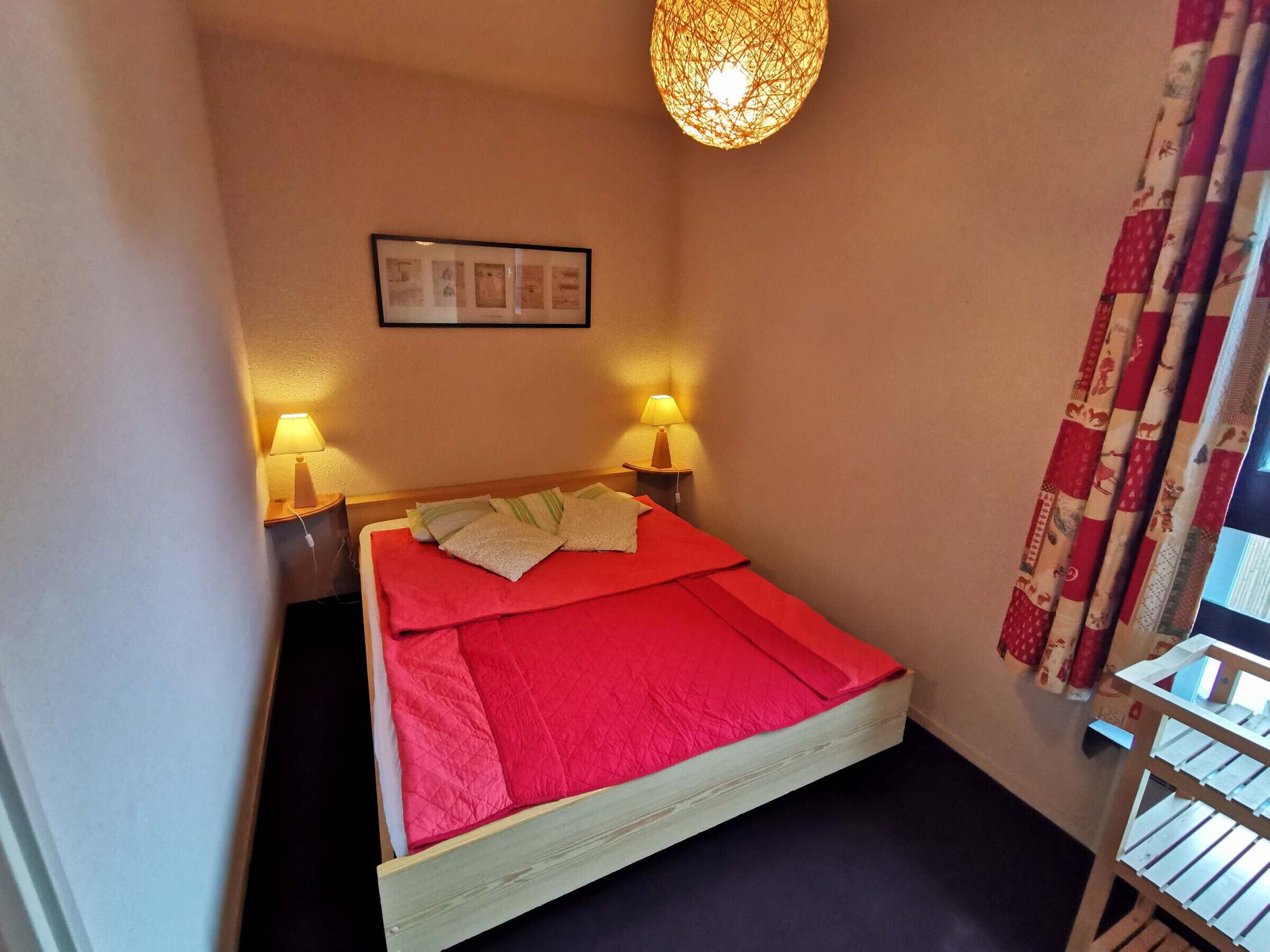 A small, cozy room with a single bed, two bedside lamps, a framed picture on the wall, and a window with curtains.