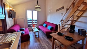 A cozy room with a kitchenette, a wooden dining table, a red sofa, a wooden staircase, and a large window with a view. - Cozy apartment for 4-6 with balcony near slopes, TV, elevator and ski locker (Les Adrets-Prapoutel)