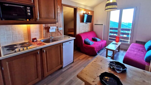 A compact kitchen with wooden cabinets, a microwave, and a sink. There's a red sofa with cushions, a small table with glasses, and a hanging light fixture. - Cozy apartment for 4-6 with balcony near slopes, TV, elevator and ski locker (Les Adrets-Prapoutel)