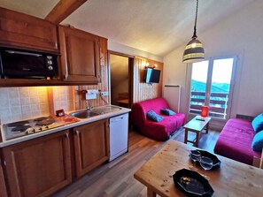 Cocina compacta con armarios de madera, microondas y un lavabo. Hay un sofá rojo con cojines, una mesita con vasos y una lámpara colgante.