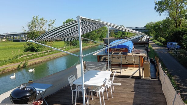 Une terrasse avec une table et des chaises, un barbecue et un espace abrité offrant une vue sur un canal et des cygnes.