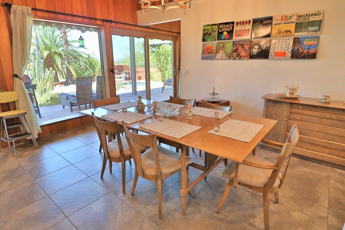 A dining area with a wooden table and chairs, a kitchen counter, and a view of the outdoors through large windows.