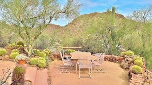 An outdoor patio with a wooden table and chairs, surrounded by cacti and desert vegetation. - Desert Paradise at Trails End (Tucson)