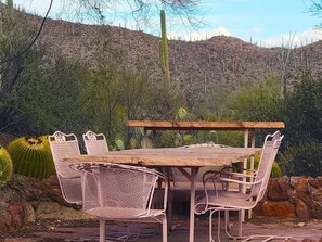 An outdoor dining area with a wooden table and wicker chairs, surrounded by cacti and a rocky hillside. - Desert Paradise at Trails End (Tucson)