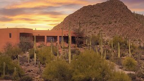 A desert landscape with a building featuring a curved facade and large windows, surrounded by cacti and a hill in the background. - Desert Paradise at Trails End (Tucson)