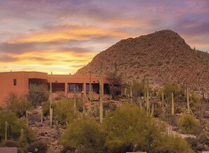 A desert landscape with a building featuring a curved facade and large windows, surrounded by cacti and a hill in the background.