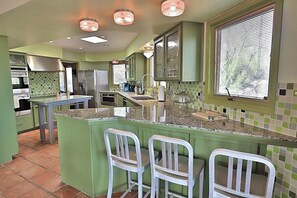 Une cuisine avec des meubles de couleur verte, un plan de travail en granit et une fenêtre avec des stores.