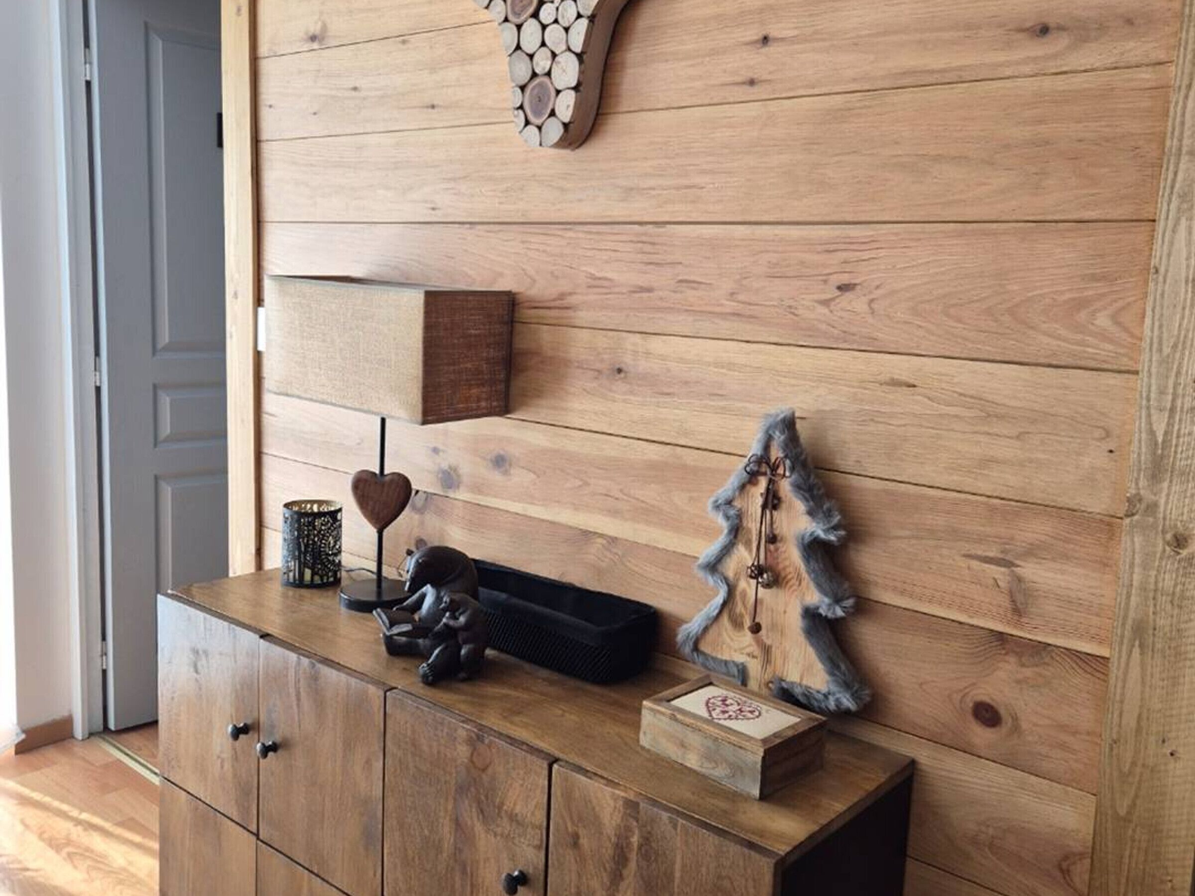 Een houten kastje met een kaarshouder in de vorm van een hart, een klein beeldje en een houten kerstboomversiering, tegen een muur met een houten paneelpatroon.