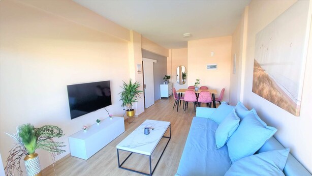 A modern living room with a flat-screen TV, a sofa, a coffee table, and a dining area with pink chairs and a wooden table. - The Urban Oasis: Comfort 2br near the City Center! (Larnaca)