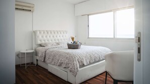Une chambre à coucher avec un lit blanc, une table de chevet, une chaise et une fenêtre.