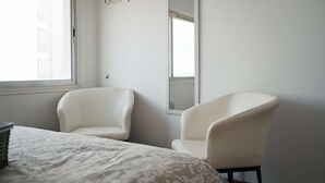 A bedroom with two white chairs, a bed with a patterned cover, and a window with a view of buildings. - Cozy Sea View Apt w/ pool access + Netflix (Larnaka)