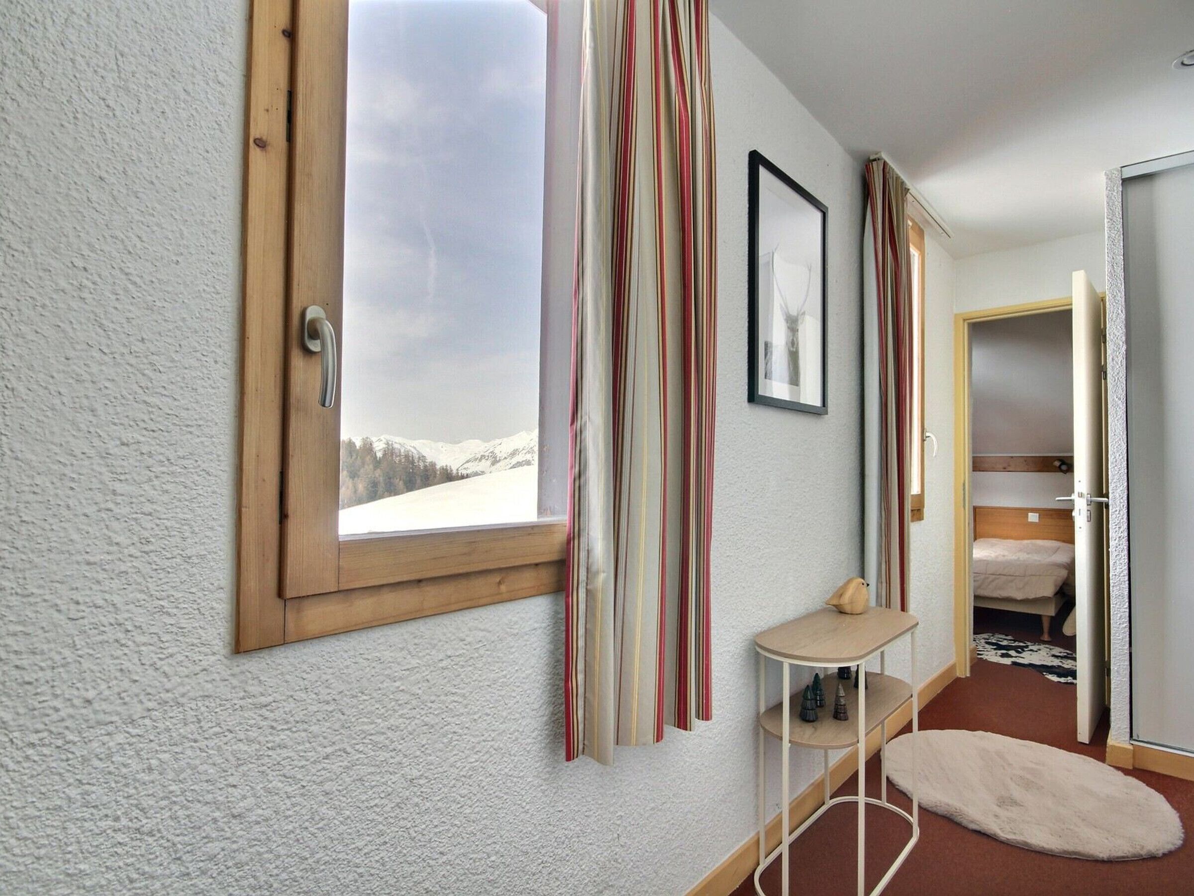 A room with a large window, a bed, a small table, and a shelf with decorative items.