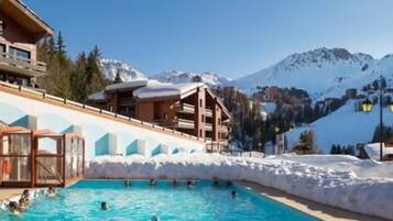 A pool area with people swimming, surrounded by buildings and snow-covered mountains.