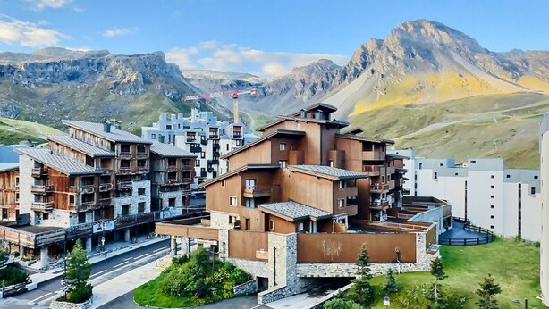 A modern hotel with multiple buildings, featuring a mix of wooden and concrete structures, set against a backdrop of mountains and clear skies.