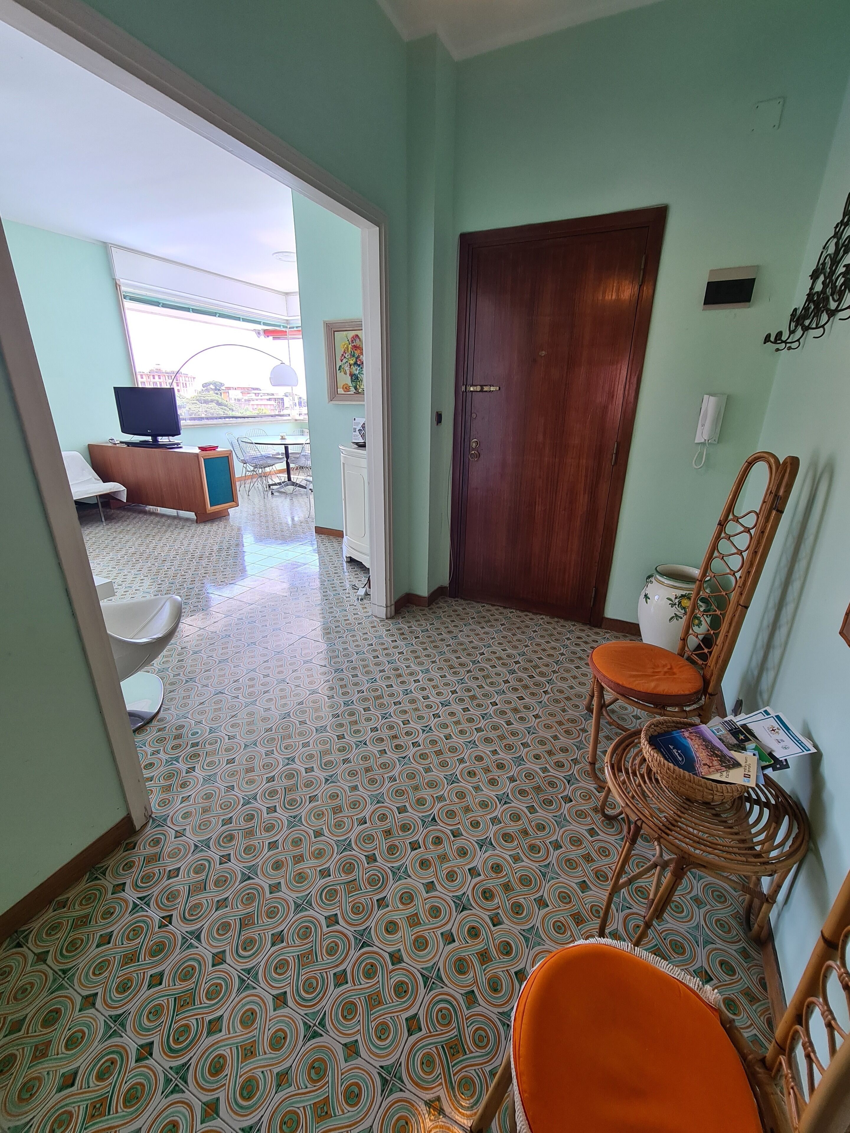 A room with patterned floor tiles, a wooden door, and a wicker chair with an orange cushion.