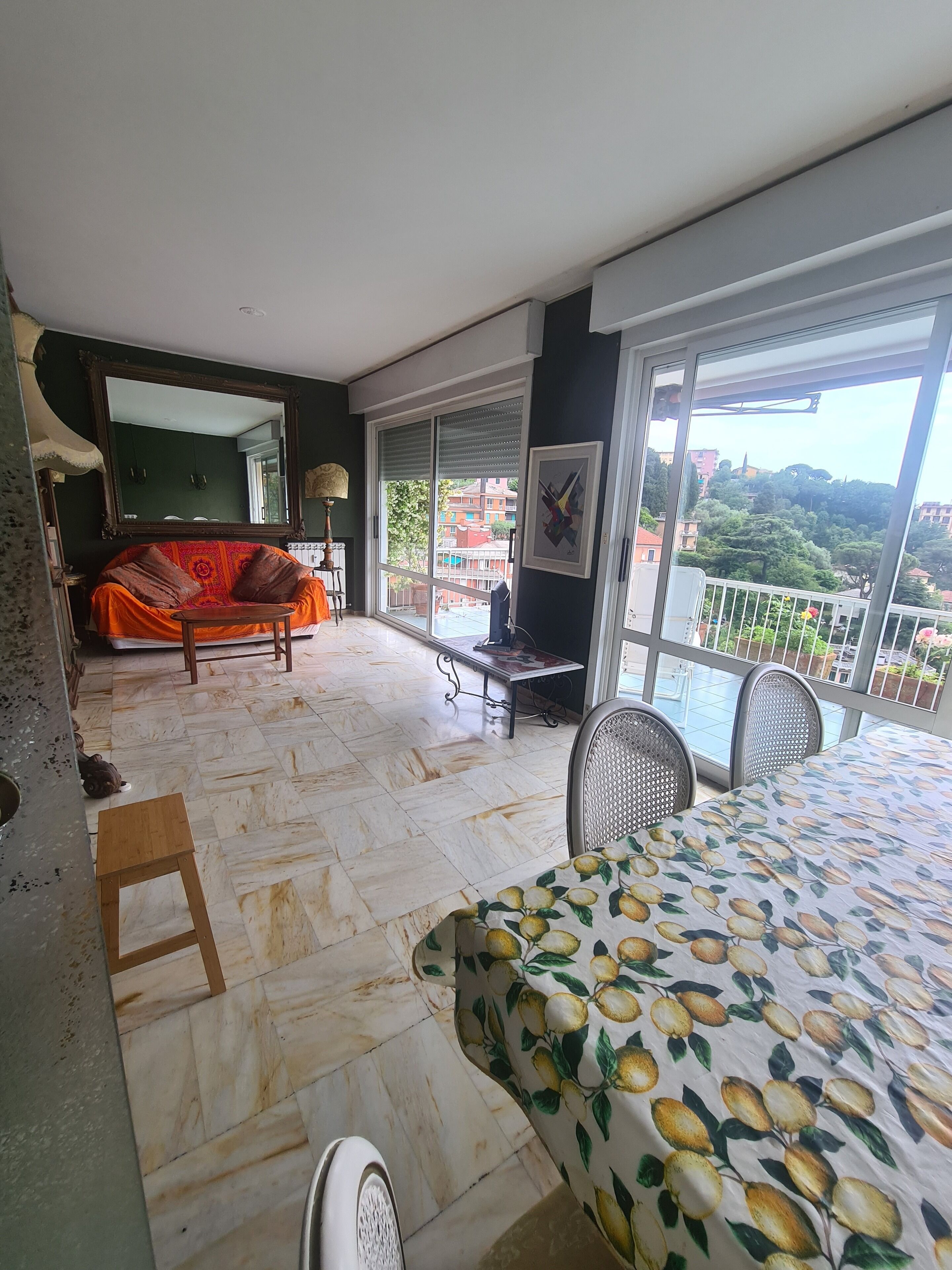 A balcony with a view of buildings and trees, a dining area with a table and chairs, and a sofa with a colorful blanket.