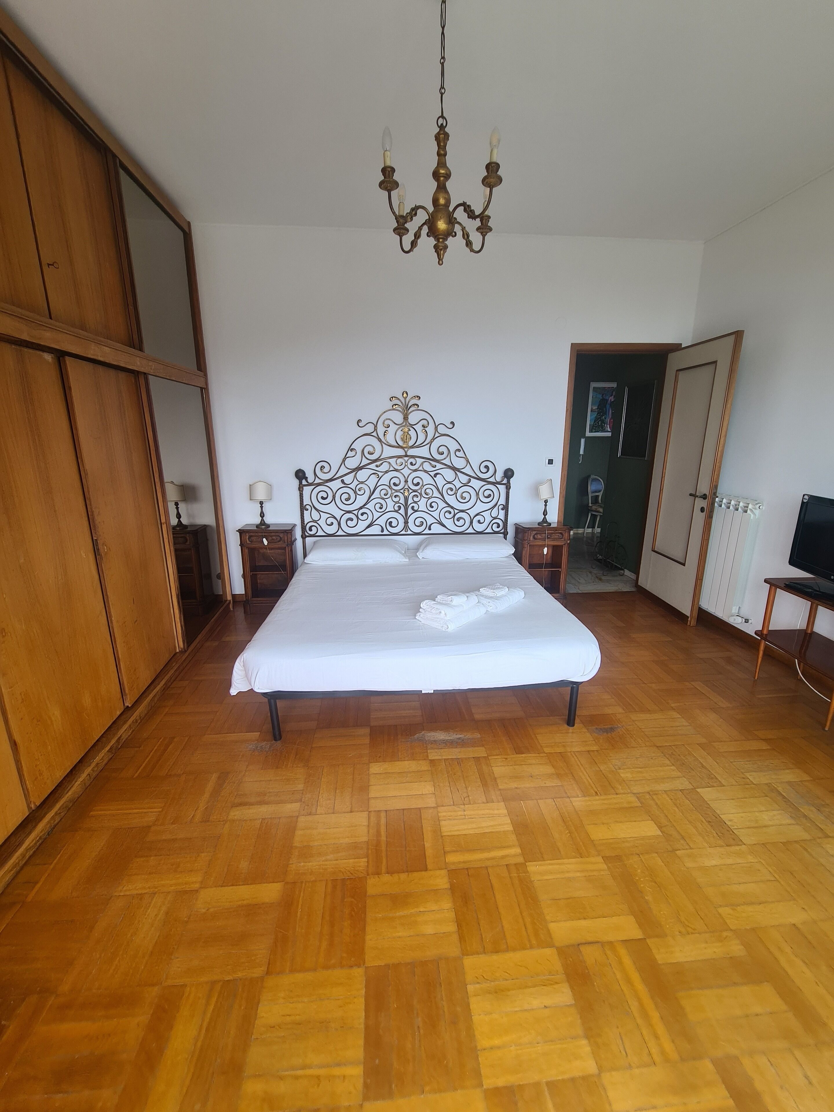 Een slaapkamer met een houten vloer, een bed met een wit dekbed, een kroonluchter, houten kasten en een televisie op een houten dressoir.