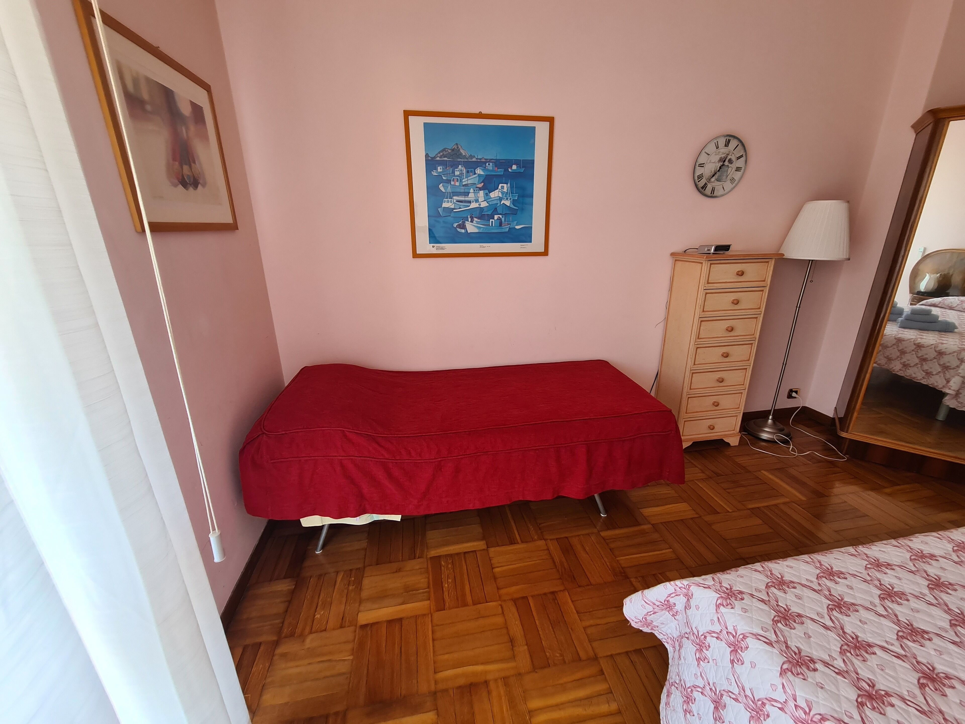 Un dormitorio con una cama roja, suelo de madera, un tocador, una lámpara, un reloj y un cuadro en la pared.