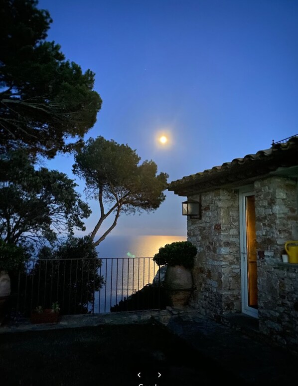 A stone building with a balcony overlooking a body of water, a tree in the foreground, and a moon in the sky.