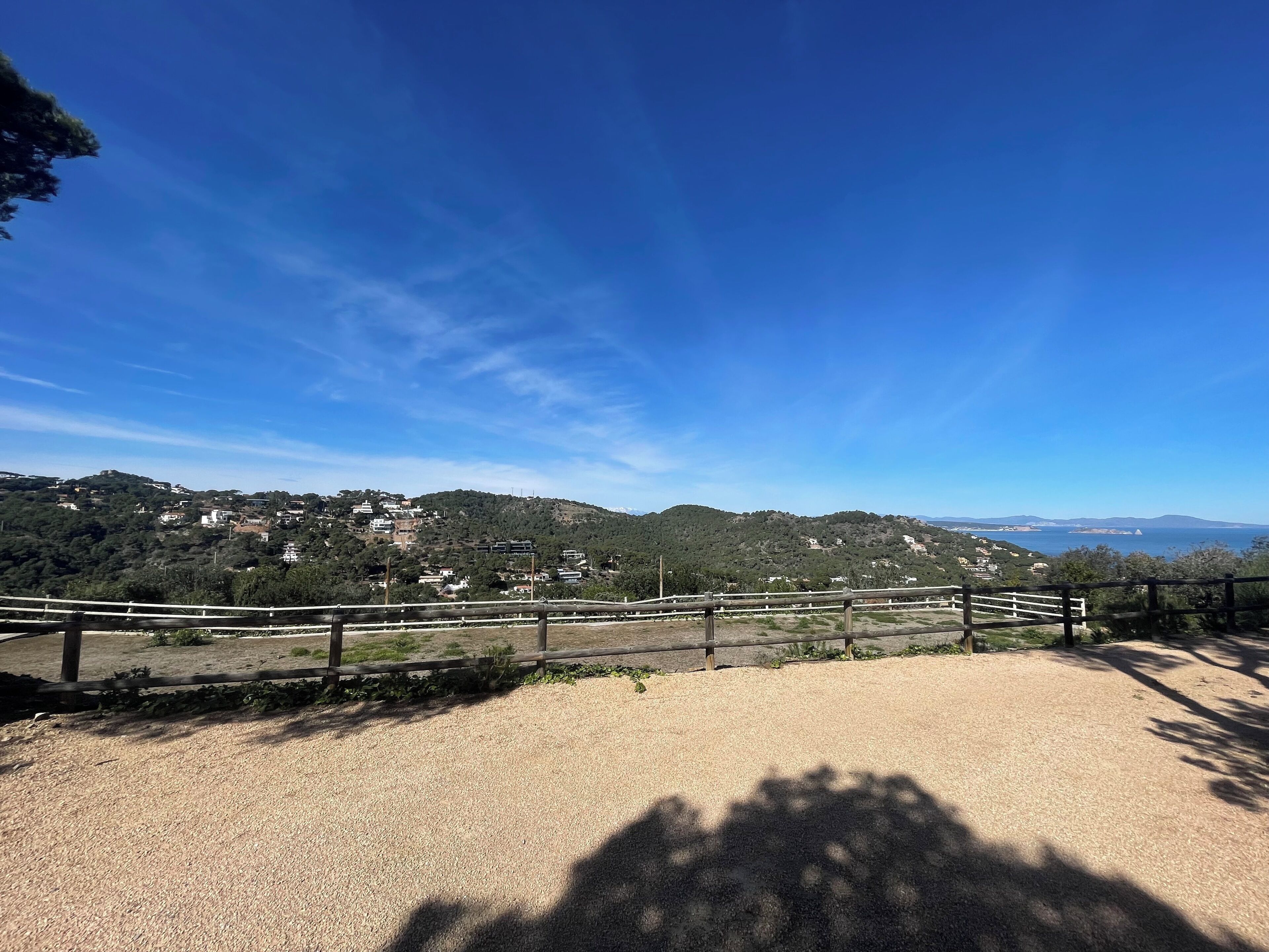 A gravel path with a wooden fence, leading to a scenic overlook with a view of a hillside village and distant mountains under a clear blue sky.