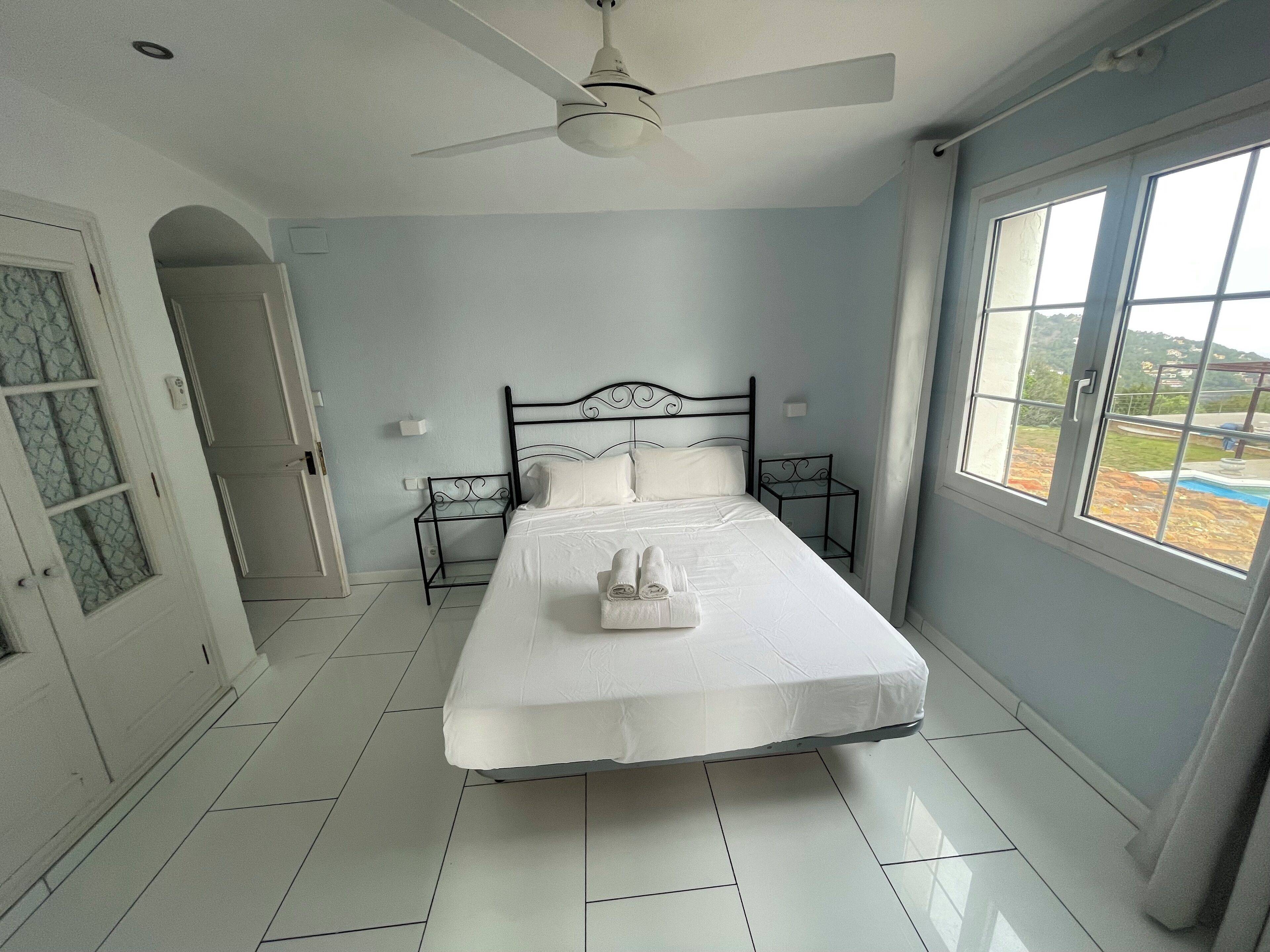 A neatly arranged bedroom with a bed, a ceiling fan, a window with a view, and a door with a frosted glass panel.