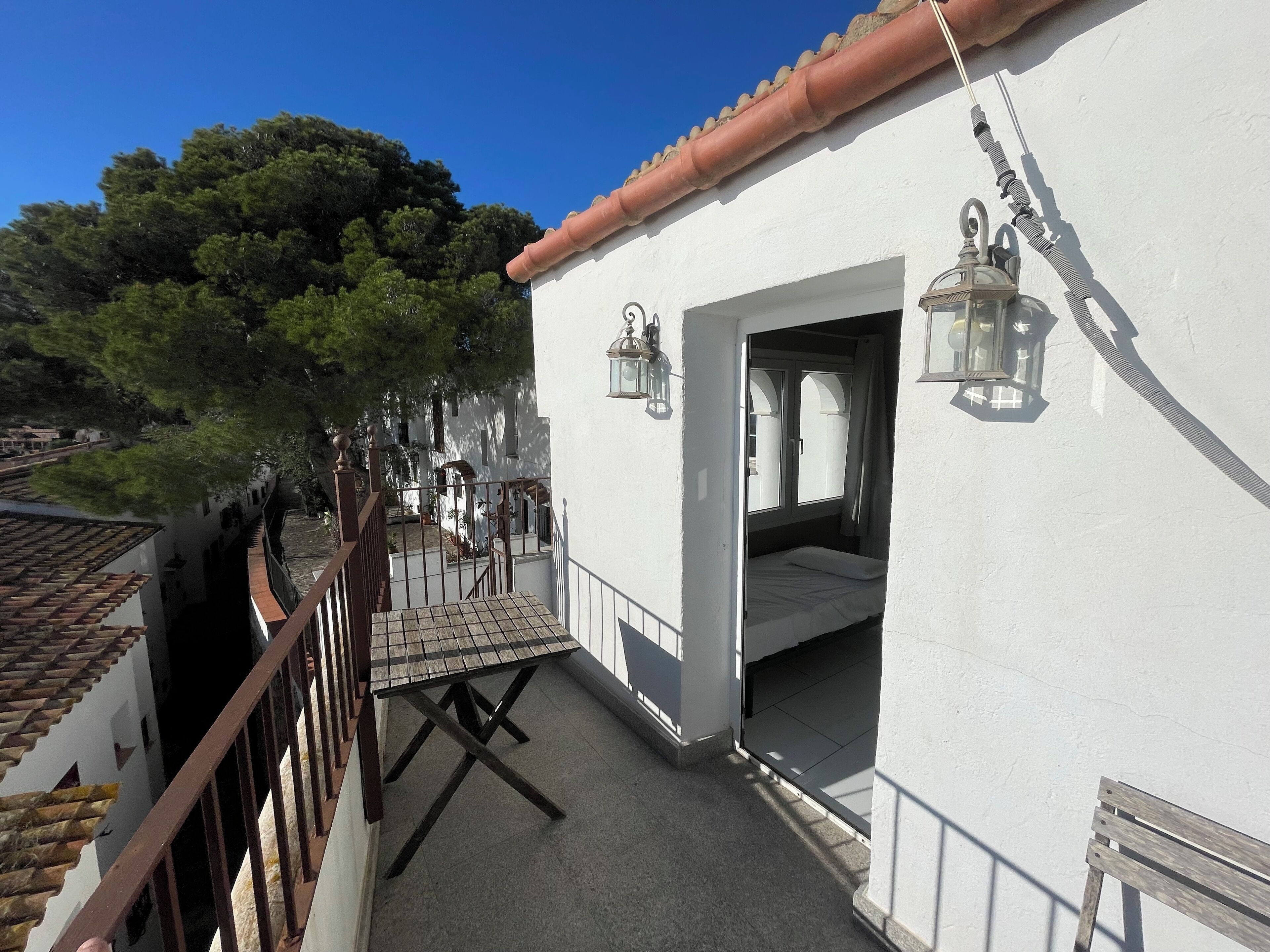 A white building with a tiled roof, a balcony with a wooden table, and two wall-mounted lanterns.