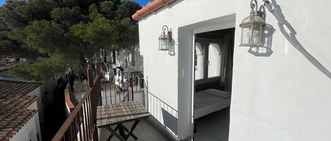 A white building with a tiled roof, a balcony with a wooden table, and two wall-mounted lanterns.