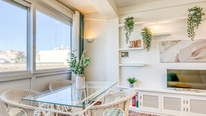 Ein modernes Wohnzimmer mit einem Glastisch, umgeben von Korbstühlen, einem weißen Schrank mit Regalen und hängenden Pflanzen.