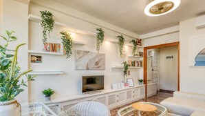 Ein modernes Wohnzimmer mit Fernseher, weißen Korbmöbeln und Pflanzen auf Regalen.