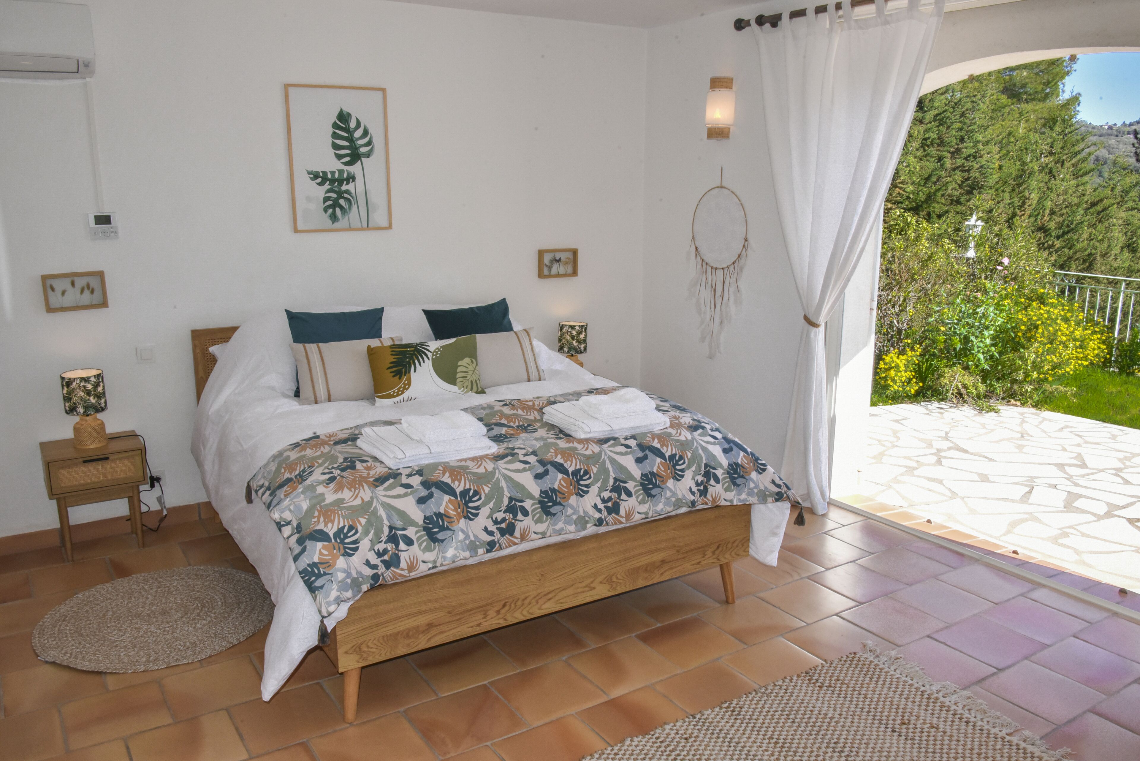 Ein Schlafzimmer mit einem Bett, einem Nachttisch, einer Wand-Klimaanlage, einem Blatt-Kunstwerk im Bilderrahmen, einem Traumfänger und Blick auf einen Garten durch ein Fenster.