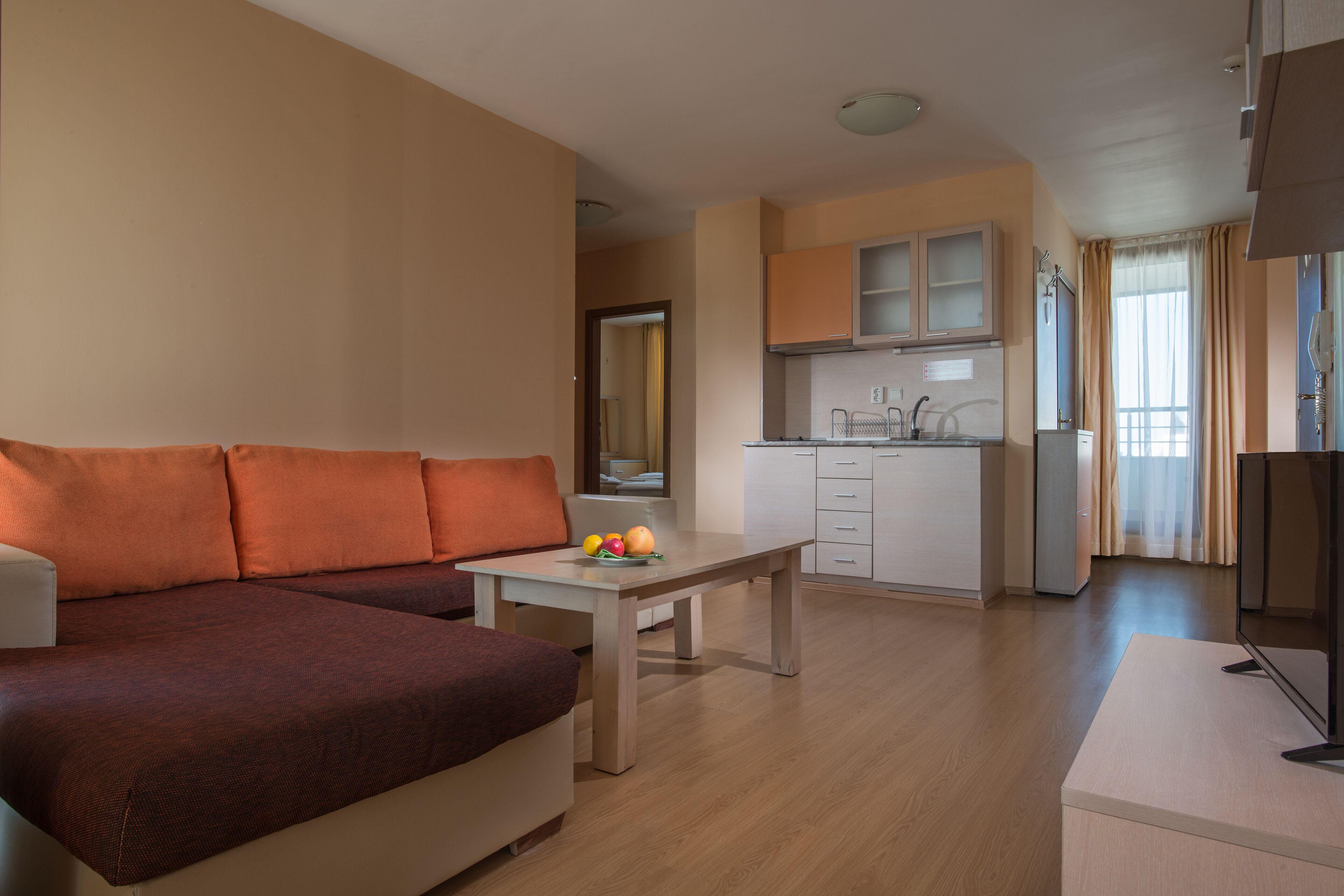 A modern living room with a sofa, a dining table with fruit, a kitchen area with cabinets, and a TV.