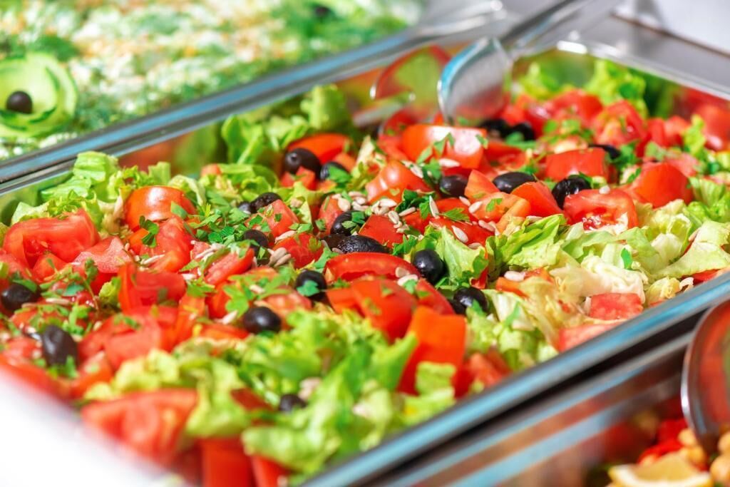 A buffet with various salads, including one with tomatoes, lettuce, and black olives.
