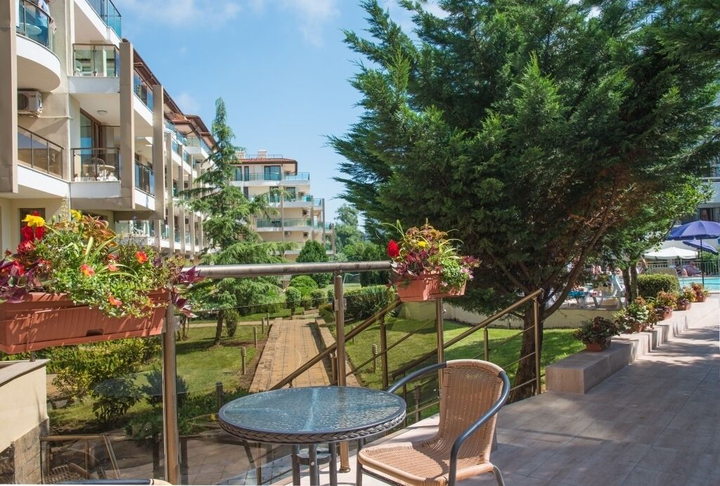 A balcony with a glass table and wicker chair, overlooking a pool and a garden with potted plants.