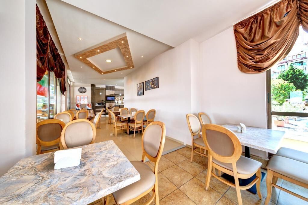 A restaurant with marble-topped tables and wooden chairs, a kitchen in the background, and large windows with curtains.