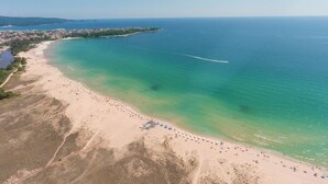 An aerial view of a coastal area with a sandy beach, clear turquoise waters, and a distant shoreline with buildings. - PRESTIGE CITY2  (Primorsko)