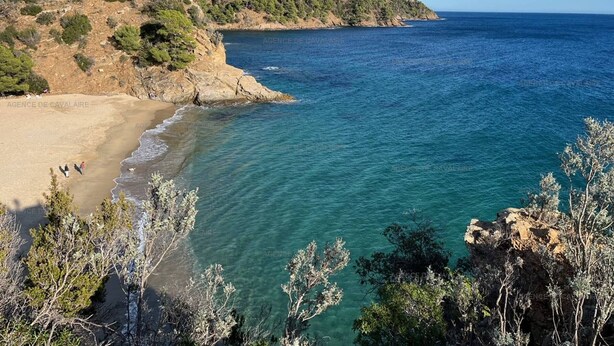 A coastal landscape with a sandy beach, clear blue sea, and rocky cliffs.