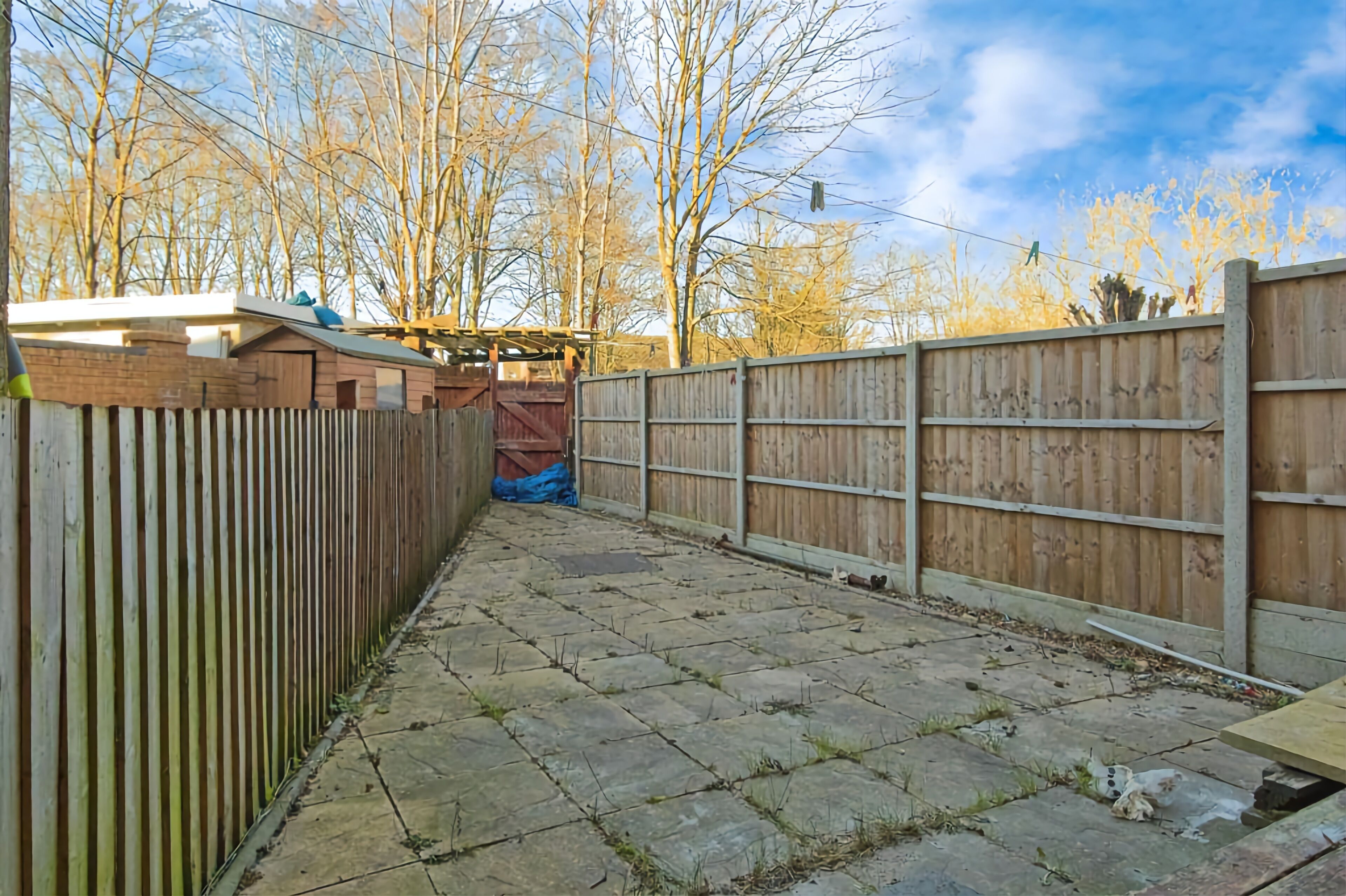 A paved backyard with a wooden fence, a covered structure, and trees in the background.