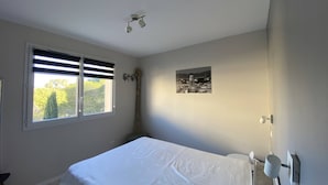 A bedroom with a bed, a window with blinds, a wall-mounted picture, and a ceiling light fixture. - Modern T3 in Central Cavalaire - Walk to Beach & Shops, Parking, A/C, Spacious Terrace (Cavalaire-sur-Mer)