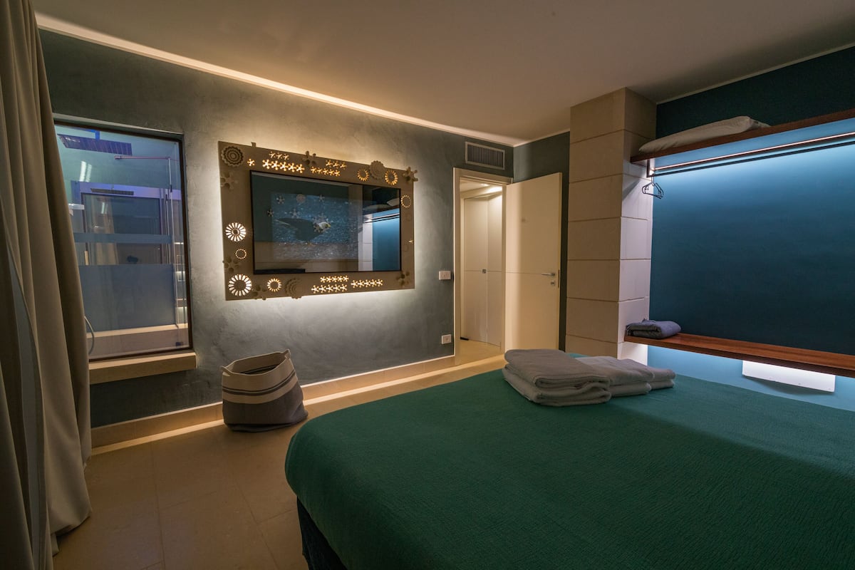 Ein Hotelzimmer mit einem großen Bett, einem Spiegel mit dekorativer Beleuchtung, einem Fenster mit Ausblick und einer Tür zu einem anderen Raum.