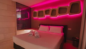 A modern hotel room with a large bed, pink ambient lighting, and a unique wall design.