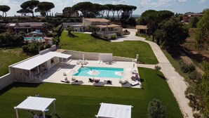 An aerial view of a resort with a swimming pool surrounded by lounge chairs and umbrellas, a well-maintained lawn, and a variety of buildings in the background.
