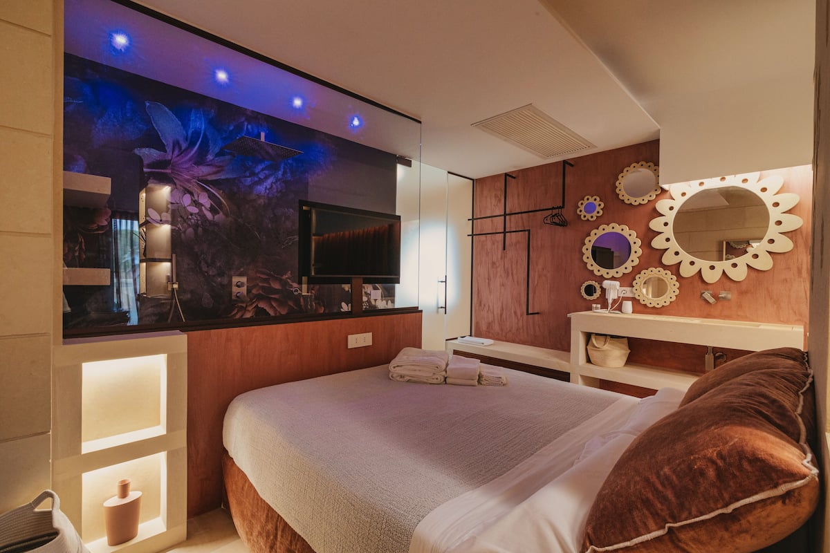 Ein modernes Schlafzimmer mit einem großen Bett, einem Schminktisch mit Spiegel und einem an der Wand befestigten Fernseher.