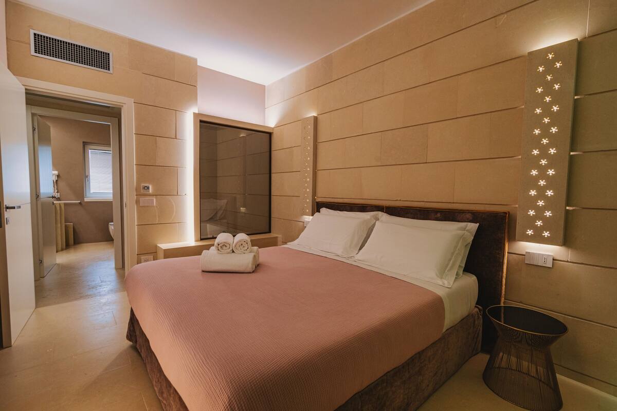 Ein modernes Hotelzimmer mit einem großen Bett, einem Nachttisch, einer Wandleuchte und Blick auf den Flur.