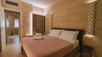 A modern hotel room with a large bed, a bedside table, a wall-mounted light fixture, and a view of the hallway.