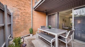 A patio with a table and chairs, a potted plant, and a sliding glass door leading to an indoor space. - GP46A | G. Prairie Haven | 2BD | Pool | Patio (Grand Prairie)