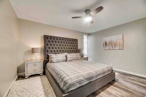 Een slaapkamer met een gebostel hoofdeinde, een bed met gestreepte beddengoed, een nachtkastje met een lamp, en een plafondventilator.